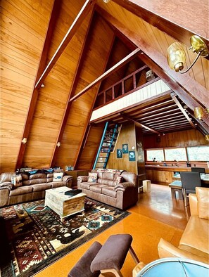 A spacious loft with high ceilings, wooden beams, a large sofa, a coffee table, and a staircase leading to an upper level.