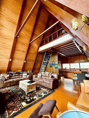 A spacious loft with high ceilings, wooden beams, a large sofa, a coffee table, and a staircase leading to an upper level.