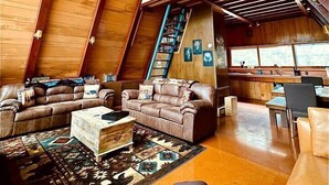 A spacious loft with high ceilings, wooden beams, a large sofa, a coffee table, and a staircase leading to an upper level. - Spacious Well Furnished A-Frame with Sauna in Union Dale, Pennsylvania (Union Dale)