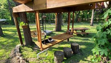 A wooden pergola structure with a bench and a table, surrounded by trees and grass.