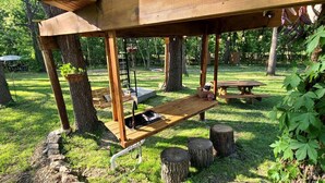 Une structure en bois formant une pergola, avec un banc et une table, entourée d’arbres et d’herbe.