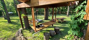 Een houten pergola met een bankje en een tafel, omgeven door bomen en gras.