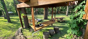 A wooden pergola structure with a bench and a table, surrounded by trees and grass.