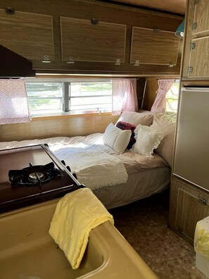 A compact RV interior with a bed, a small kitchenette, and a window providing natural light.