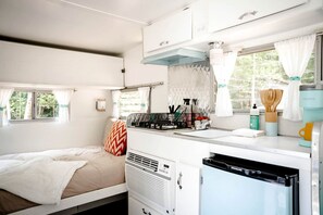 A compact RV interior with a bed, kitchenette, and a window with curtains.
