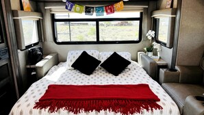 Een slaapkamer met een bed, kussens, een rode deken, een raam, kasten en een klein tafeltje met een plant.