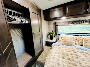 A bedroom with a bed, a closet with hanging clothes, a nightstand with a plant, and a window with a view. - Spacious Camper on a Llama Rescue Ranch with Stunning Views near Questa, New Mexico (Questa)