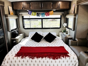 Ein Schlafzimmer mit Bett, Kissen, einer roten Decke, einem Fenster, Schränken und einem kleinen Tisch mit einer Pflanze.