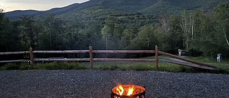 A gravel area with a fire pit, a wooden fence, and a view of a forested mountain range.