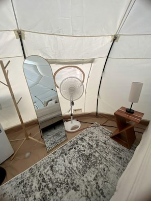 Tienda de campaña con cama, ventilador, espejo y una mesita de noche con lámpara.