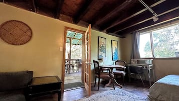 A room with a wooden ceiling, a dining table with chairs, a bed, a desk with a chair, and a window.