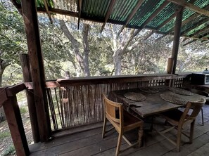 An outdoor wooden deck with a table and chairs, surrounded by a bamboo fence, and a covered area with a corrugated roof.