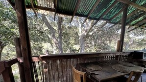 An outdoor wooden deck with a table and chairs, surrounded by a bamboo fence, and a covered area with a corrugated roof.