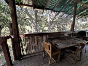 Una terraza de madera al aire libre con una mesa y sillas, rodeada por una cerca de bambú y un área cubierta con techo de láminas corrugadas.