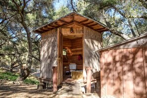 Una cabaña de madera con techo inclinado, un pequeño porche y un interior visible con una cama y un ventanal.