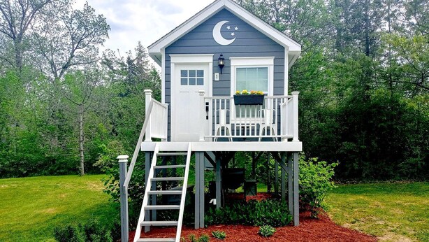 A two-story house with a balcony, a ladder leading to the upper level, and a small garden area with mulch and plants. - Beautiful Treehouse Under The Stars Near Wolverine, Michigan (Holly)