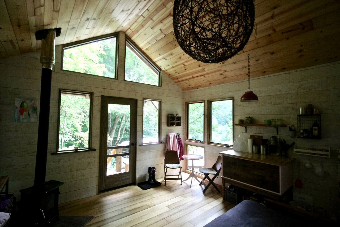 L’intérieur d’un chalet rustique, avec un grand poêle en bois, de nombreuses fenêtres, un luminaire suspendu et un coin repas.