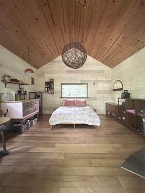 A cozy cabin-style bedroom with a wooden ceiling, a large bed with a patterned bedspread, a wooden dresser, and a hanging light fixture. - Off Grid Tiny Cabin Ideal for Couples in Burnsville, North Carolina (Burnsville)