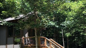 A wooden deck with a staircase leading to a treehouse, surrounded by dense foliage. - Off Grid Tiny Cabin Ideal for Couples in Burnsville, North Carolina (Burnsville)
