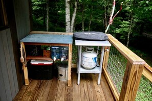 A wooden deck with a small table, a trash can, and a propane tank. - Off Grid Tiny Cabin Ideal for Couples in Burnsville, North Carolina (Burnsville)