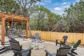 A backyard with a wooden pergola, hanging chairs, Adirondack chairs, and a stone fire pit.
