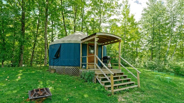 A yurt with a wooden staircase leading to a covered entrance, surrounded by a grassy area and trees. - Beautiful Upstate New York Luxury Camping Yurt on a Farm (Granville)