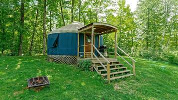 A yurt with a wooden staircase leading to a covered entrance, surrounded by a grassy area and trees.