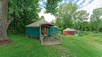 A yurt with a wooden porch and a small deck, surrounded by lush greenery and other yurts in a campsite.