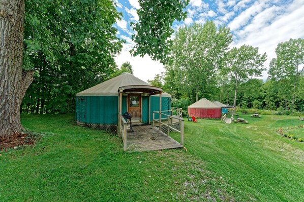 A yurt with a wooden porch and a small deck, surrounded by lush greenery and other yurts in a campsite.