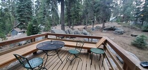 Eine Holzterrasse mit einem runden Tisch und vier Stühlen, umgeben von bewaldetem Gelände mit großen Felsen und einer kleinen Hütte im Hintergrund.