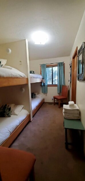A room with bunk beds, a chair, a table with a stack of towels, a window with curtains, and a mirror on the wall.
