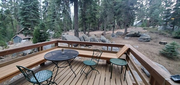A wooden deck with a round table and four chairs, surrounded by a forested area with large rocks and a small cabin in the background.
