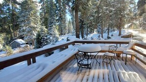 Una terraza de madera con suelo cubierto de nieve, rodeada de árboles cargados de nieve y mobiliario exterior.