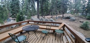Une terrasse en bois avec une table ronde et quatre chaises, entourée d’une zone boisée avec de gros rochers et un petit chalet en arrière-plan.