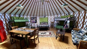 A yurt interior with a wooden floor, a dining table with chairs, a sofa, a bed with green curtains, and a ceiling fan. - Colorful Upstate New York Yurt Rental on a Farm (Granville)