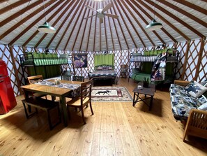 A yurt interior with a wooden floor, a dining table with chairs, a sofa, a bed with green curtains, and a ceiling fan. - Colorful Upstate New York Yurt Rental on a Farm (Granville)