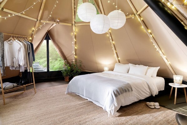 A tented accommodation room with a bed, hanging clothes rack, potted plant, and string lights.