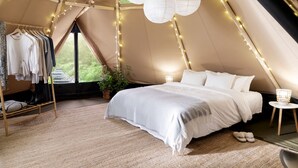 Quarto em alojamento tipo tenda, com cama, cabideiro, planta em vaso e luzes de cordão.