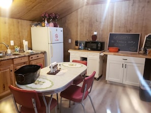 A kitchen with wooden walls, a white refrigerator, a microwave, a chalkboard with a welcome message, and a table with chairs.