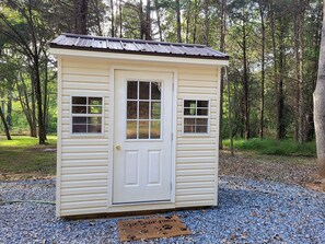 A small, single-story shed with a door and two windows, surrounded by trees and gravel. - Private Pet-Friendly Cabins with Fire Pit for Nature Escapes in Clear Spring, Maryland, for Memorable Family Glamping Trips (Clear Spring)