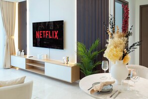 A modern living room with a flat-screen TV displaying 'NETFLIX', a white sofa, a dining table set with a vase of flowers, and a minimalist console table.