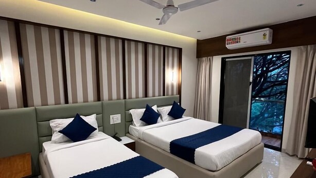 A hotel room with two beds, a ceiling fan, a large window with curtains, and a flat-screen TV. - Hotel Sai Residency (Pune)