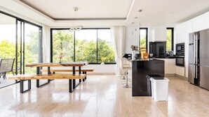 A modern kitchen with a large island, a dining area with wooden benches, and large windows offering a view of greenery. - Astoria Villa #2 – 3 Bed Pool Villa in Koh Samui (Chang Wat Surat Thani)