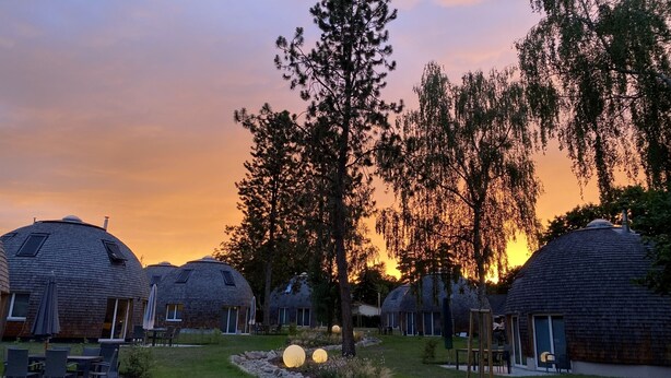 Fileira de cabanas em formato de cúpula com áreas de estar ao ar livre, rodeadas por árvores e um pôr do sol ao fundo.