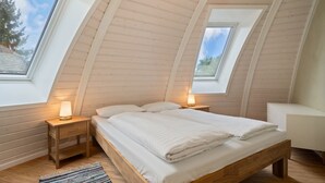 Um quarto com teto inclinado, paredes de madeira, uma cama com roupa de cama branca, duas mesas de cabeceira com luminárias e vista para árvores através de claraboias.