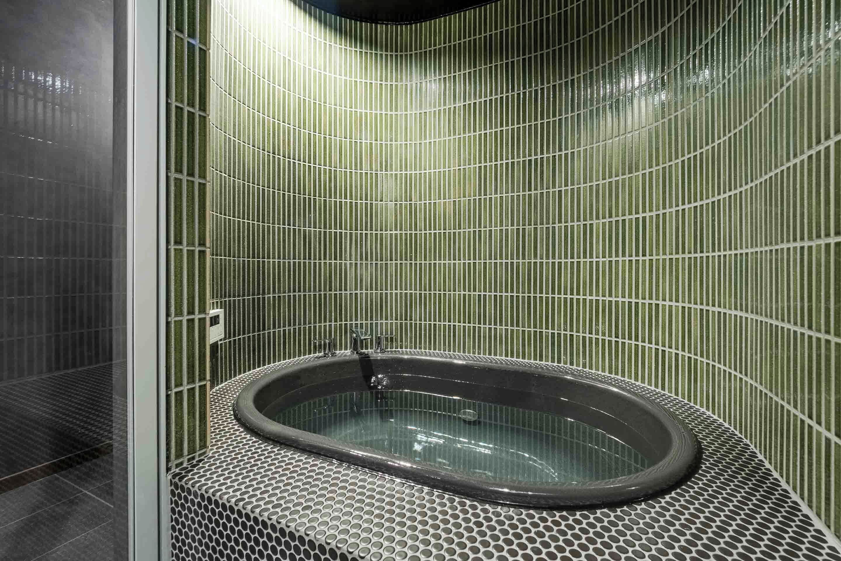 A modern bathroom with a freestanding bathtub, green vertical tiles, and a perforated metal floor.