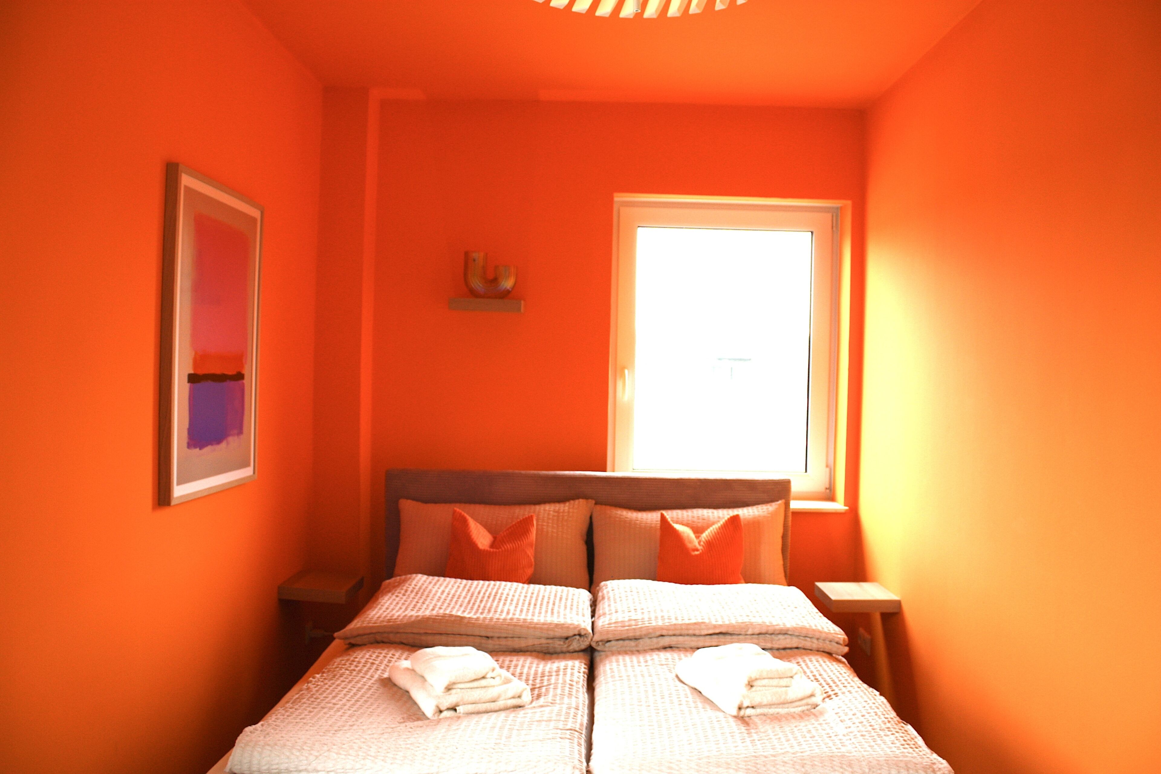 A room with orange walls, a bed with two pillows, a framed picture on the wall, and a window with blinds.