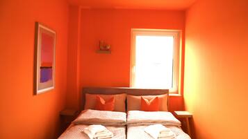 A room with orange walls, a bed with two pillows, a framed picture on the wall, and a window with blinds.