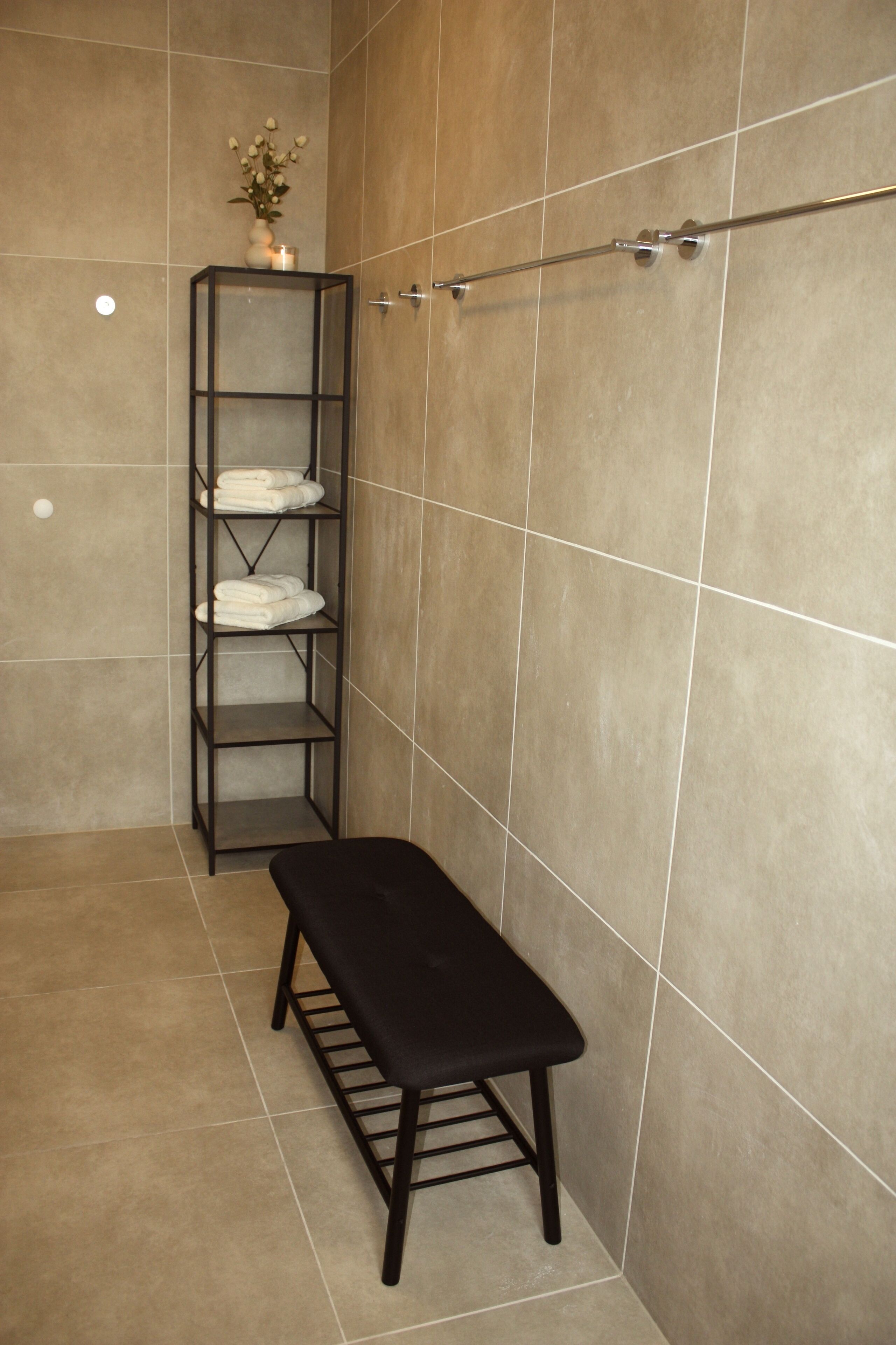 A bathroom with beige tiles, a black bench, and a metal shelf with folded towels.