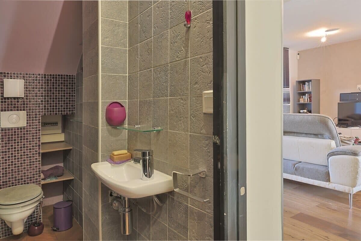 A bathroom with a toilet, sink, and shower area. There is a glass shelf with toiletries and a pink soap dispenser. The room has tiled walls and a wooden floor.